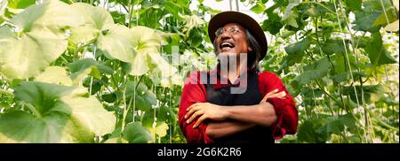 Senior middle aged male farmer having arms crossed with happy teethy smile wearing a straw hat in red farming uniform inside farm garden in summer Stock Photo