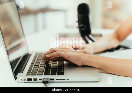 Closeup of hands of young female podcaster writing new blog for social media while typing on laptop on desk with mic in background at home Stock Photo