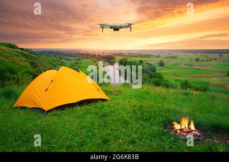 Orange tent on a hill above river under dramatic sky with quadcopter in air. Dawn over a camping and burning bonfire