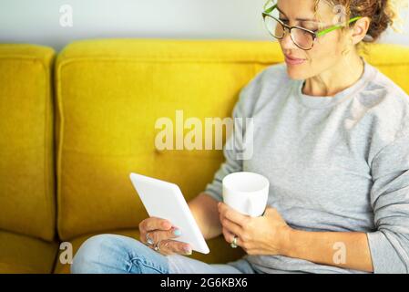 Close up portrait of mature pretty woman reading a book on tablet with glasses sitting on the yellow couch at home in break leisure time activity alon Stock Photo