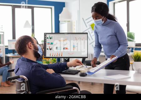 African manager with face mask working with handicapped invalid immobilized colleague in new normal business office during covid19 global pandemic, checking statistics on clipboard. Stock Photo