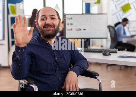Immobilized entrepreneur sitting in wheelchair waving discussing about financial strategy during video meeting on videocall. Young disabled paralized handicapped project manager talking online with partners. Stock Photo