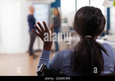 Independence handicapped african woman entrepreneur coming in start up office waving sitting immobilized in wheelchair greetting business team, diverse colleagues welcoming in startup financial company. Stock Photo