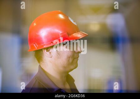 Portrait of young Asian operator worker. Red hardhat and protective glasses. Through soft-filter. Chevron plastic pipes production plant. Stock Photo