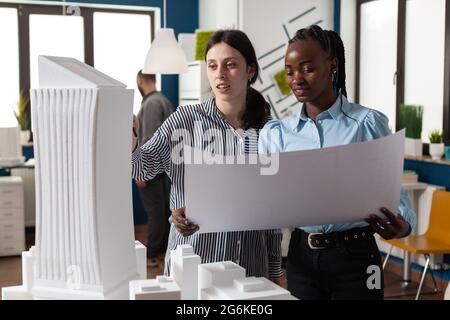 Colleagues architects women checking plans of blueprints layout design. Multi ethnic team of professional workers standing at building model maquette for modern technology project Stock Photo