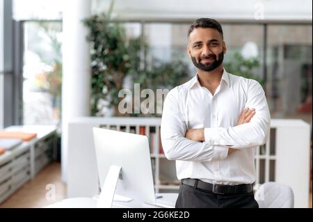 Formal business male portrait. Confident successful Indian businessman or manager, in white shirt, stands near his work desk in the office, arms crossed, looks directly at camera and smiles friendly Stock Photo