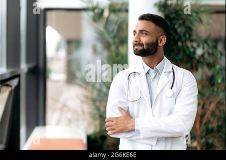 Indian handsome confident general medicine doctor, stands in a clinic, dressed in medical clothes with a stethoscope around his neck, looks away, smiling friendly. Doctor of the highest qualification Stock Photo