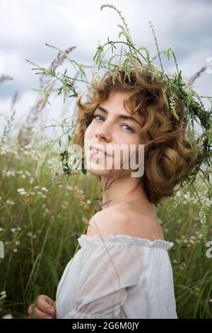 portrait of a beautiful young woman in white on a field of wild daisies in a picturesque valley. young lady 16-18 years old in wreath of flowers. Unit Stock Photo