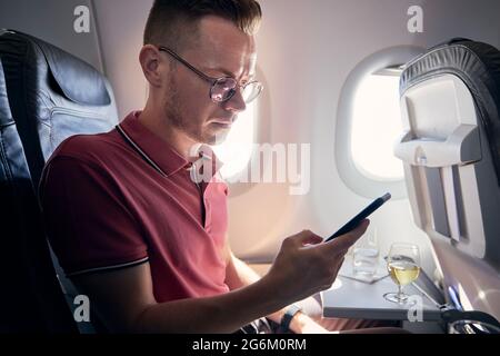 Young man using mobile phone at airplane. Passenger enjoys flight with internet connection a beverages. Stock Photo