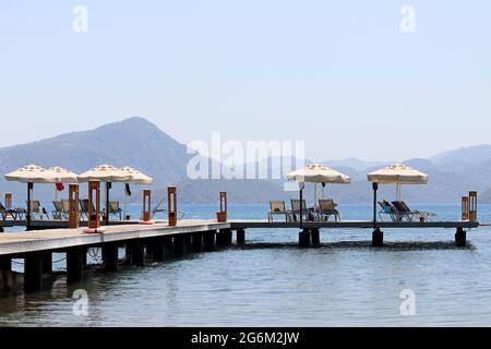 Wooden pier with lounge chairs and parasols on sea resort, vacation concept. People sunbathing on a beach on misty mountains background Stock Photo