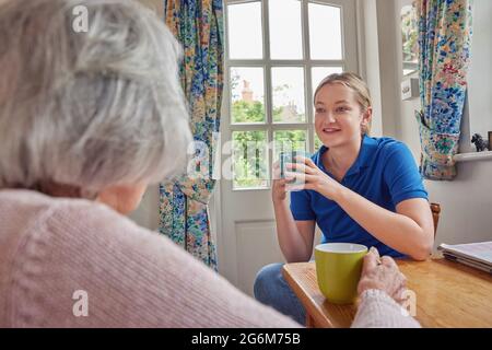 Female Home Help Having Cup Of Tea With Lonely Senior Woman In Kitchen Stock Photo