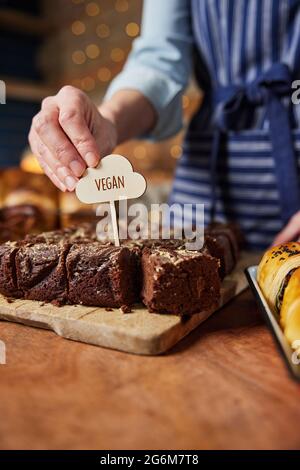 Sales Assistant In Bakery Putting Vegan Label Into Freshly Baked Chocolate Brownies Stock Photo