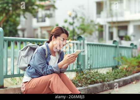 Attractive woman in city holding mobile phone, young female student with backpack looking at camera during coffee break outdoors Stock Photo