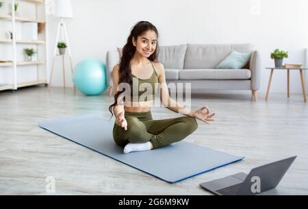 Peaceful Indian woman having online meditation or yoga class, sitting in lotus pose in front of laptop at home Stock Photo