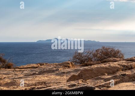 Sun rays shining though clouds over the sea rocky shore in Greece. Seascape on Mediterranean sea with island in distance in sunset light. Perspective Stock Photo