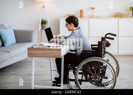 Side view of focused teen boy in wheelchair using laptop for online studies or communication with teacher at home Stock Photo