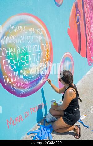 A female artist muralist writes a message about polluting the oceans with plastic on a mural on the boardwalk in Coney Island, Brooklyn, New York City. Stock Photo