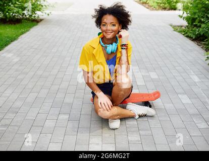 Positive teenage girl with vitiligo sitting on red skateboard outdoor at city park Stock Photo