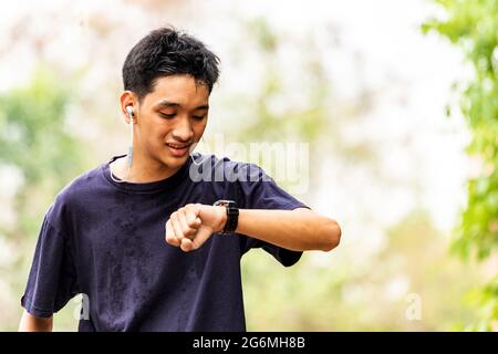 A smiling young man is jogging in the park. He is looking at his watch. He listens to music on his mobile phone, upper body approaching the camera wit Stock Photo