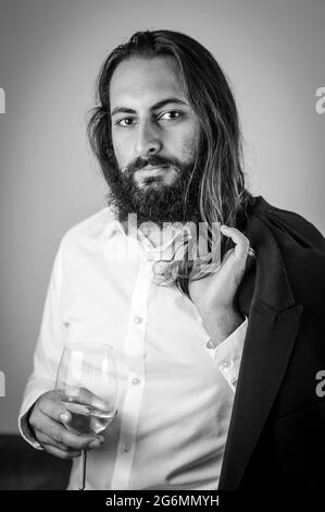 Portrait of a young middle eastern businessman with beard and long hair holding a glass of white wine and his jacket on his shoulder Stock Photo