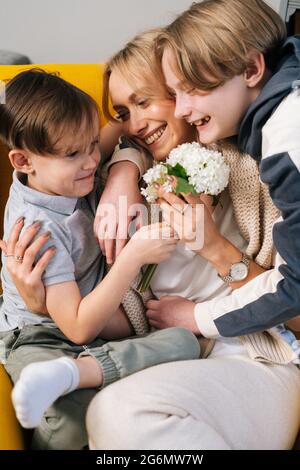 Close-up of two cheerful little funny kid child sons congratulating happy young mom with mothers day presenting bouqert of flowers.