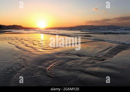 Location place Agia Marina Beach, island Crete, Greece. Sea coast spangled by rocks, the sunrise is reflecting on the wet sand. The mountains in haze Stock Photo