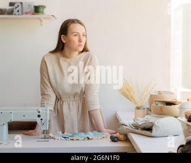 Young seamstress standing near workplace at fashion atelier and looking away with thoughtful look Stock Photo