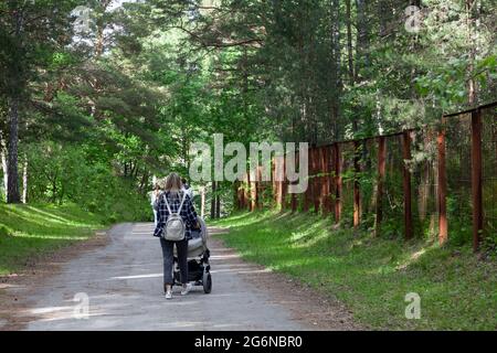 Back view of mother walking with her baby in stroller. Grey stroller outside. Young modern mom with baby son in stroller walking in Sunny Park. Concep Stock Photo