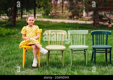 a cute, mischievous child is sitting on chairs and waiting for friends for a birthday party for an open-air holiday in the garden near the house on a Stock Photo