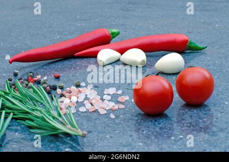 a sprig of rosemary and red chili with salt and spices on a glossy dark stone surface. Cloves of garlic and cherry tomatoes. Stock Photo