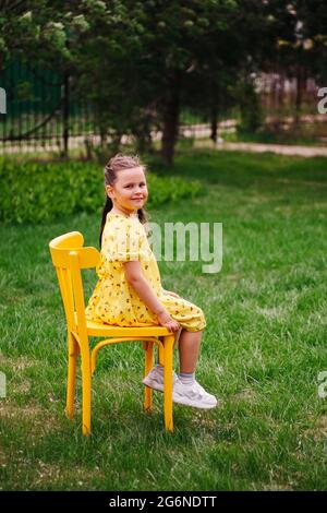 full-length portrait of a beautiful girl in a yellow dress on a yellow Viennese chair in the garden in the backyard of a village house on a sunny day Stock Photo