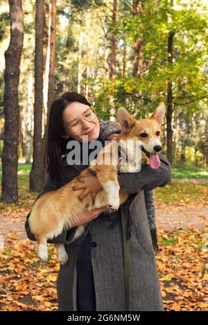 Woman in a grey coat walking with a dog in an autumn park. Cute corgi on fall foliage Stock Photo