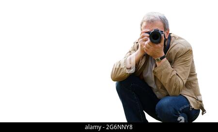 A middle-aged man with a camera takes pictures while squatting. In the foreground, the camera is aimed at the viewer. An experienced photographer, iso Stock Photo