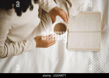 African-American woman on white bed drinking coffee and reading book. Stock Photo