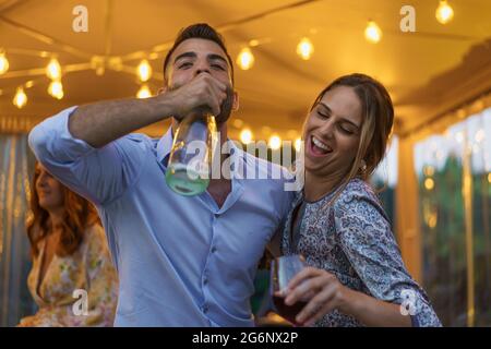 Young couple drinking sparkling white wine from the bottle during a night event party. People having fun drinking alcoholic drinks concept. Stock Photo