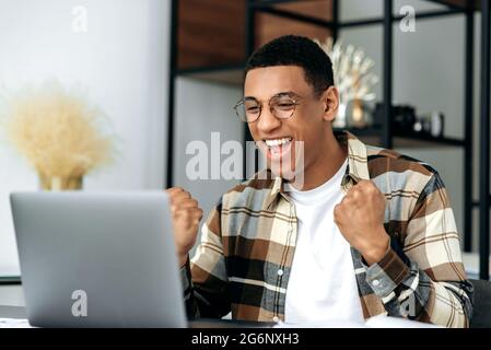Amazed excited latino male freelancer or office worker, sitting at his work desk at office, using laptop, shouts and rejoice in success, win or good profit, looking at laptop and gesturing hands Stock Photo