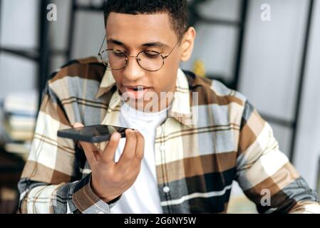Cell phone communication. Focused latino guy in casual shirt and with glasses, sits at a desk and recording an audio message or talking on smartphone on speakerphone with friend or family Stock Photo