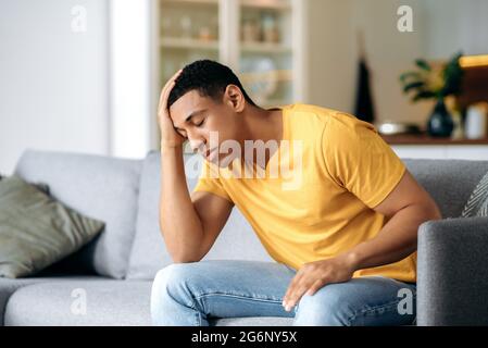 Frustrated sad handsome latino young alone man, sitting on a sofa at home, holding his forehead, having headache, depression, disappointed, closed eyes, needs psychological support and rest Stock Photo