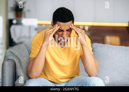 Disappointment latino guy in a yellow t-shirt sits on a sofa at home, massages his temples with his eyes closed, he has personal problems, headache, migraine, stress, thinks about solving the problem Stock Photo