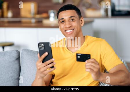 Positive attractive hispanic guy, in orange t-shirt, uses his smart phone and credit card to pay for online purchases, delivery, make an online transaction, payments, smiles happily. Shopping online Stock Photo