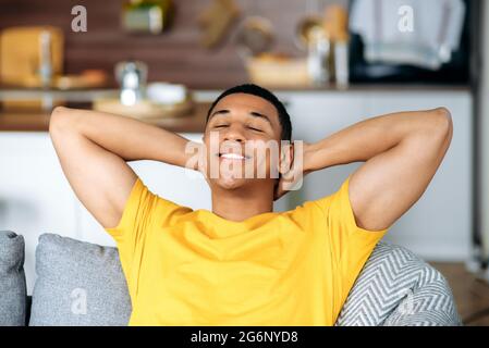 Satisfied young latino man relaxes on a sofa in living room, smiling. Handsome hispanic guy chilling at home, have lazy leisure, dreaming about vacations, throwing his hands behind head Stock Photo