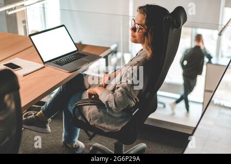 Young woman in glasses sitting in the office, thinking, laptop at her workplace. Modern communication technology, remote working and freelance job Stock Photo