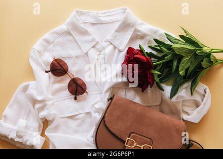 Woman fashion flat lay casual clothes with white blouse, handbag, sunglasses and dark red peony flower on beige background. Top view