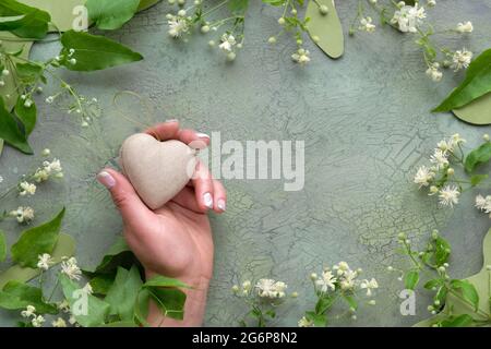 Hand holding heart from recycled craft paper. Flat lay with wild off white flowers and green leaves. Natural frame on aged textured painted green Stock Photo