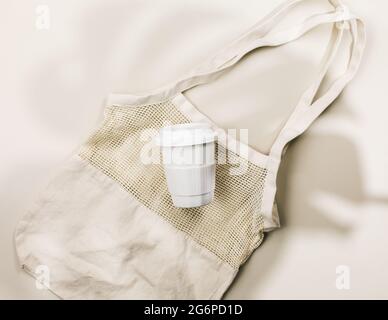 Beige reusable cotton mesh bag and coffee mug on a beige background. Eco-friendly shopping bag. Close up. Zero waste grocery concept. Sustainable life Stock Photo
