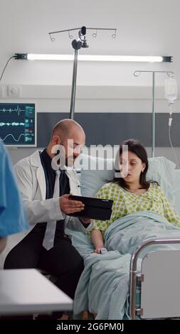 Practitioner doctor explaining disease diagnosis to sick woman using tablet computer. Patient resting in bed recovering after surgery in hospital ward while black nurse typing medical expertise Stock Photo