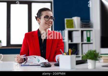 Smiling cheerful businesswoman doing paperwwork in corporate office, workplace , looking at computer display and holding charts on clipboard. Entrepreneur wearing glasses. Employee doing finance wwork. Stock Photo