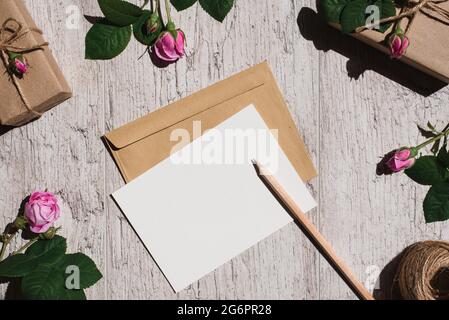 Mockup blank white greeting card or invitation, kraft brown envelope, pink rose flowers and gift boxes on gray background. Top view. Copy space Stock Photo