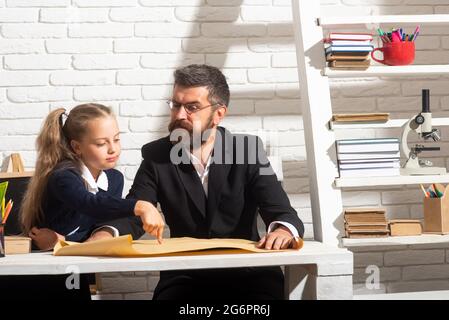 Elementary funny pupil girl with serious teacher in classroom. Fathers and daughters learning. Doing homework with dad. Stock Photo