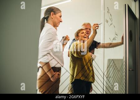 Female executive looking happy and writing on glass to explain new project to her colleagues in office. Smiling woman explaining new ideas by writing Stock Photo